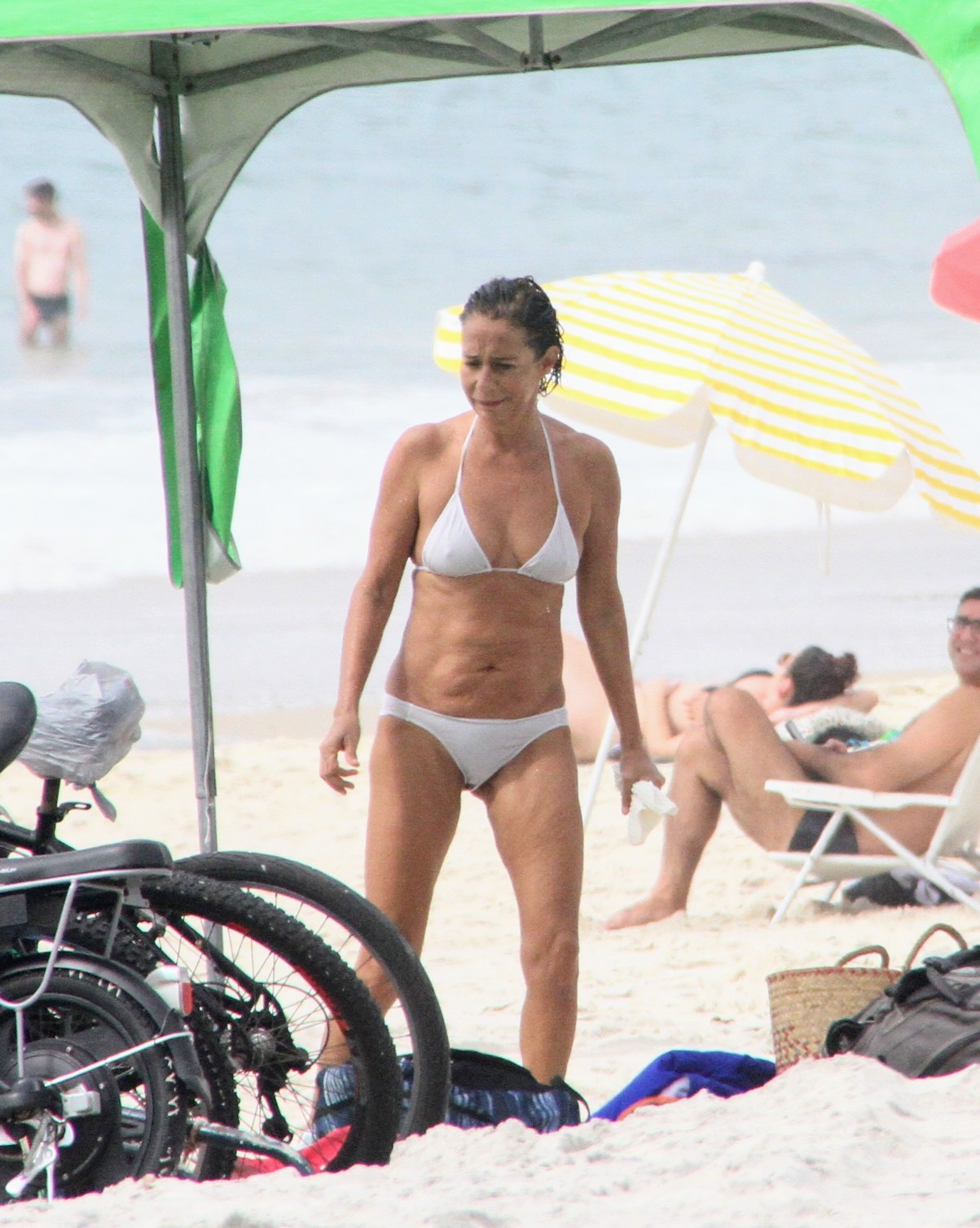 Mulher branca, de cabelo curto, usando um biquíni branco e segurando um pacote branco. Ela se encontra em meio a barracas de praia e, à sua frente, encontram-se bicicletas e bolsas