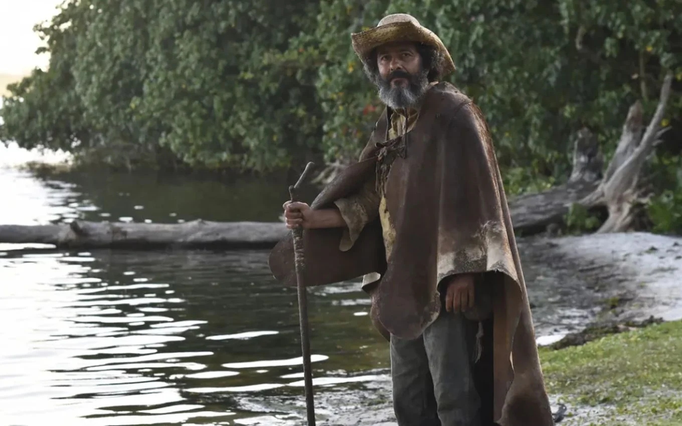 Marcos Palmeira caracterizado como a entidade Velho do Rio, da novela 'Pantanal'; ele leva um chapéu, um manto, um cajado e roupas sujas, e encontra-se à beira do rio