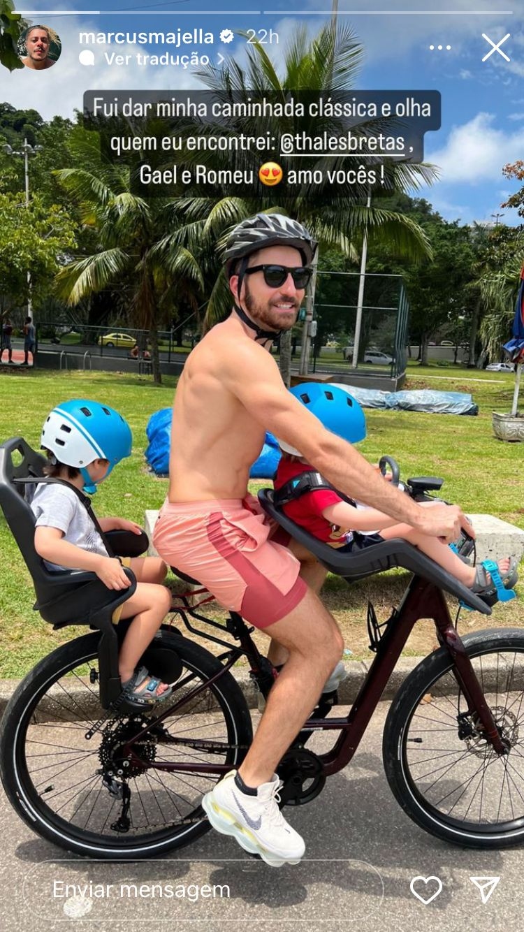 Foto de Thales Bretas sem camisa andando de bicicleta com os filhos em cadeirinhas à frente e atrás do pai